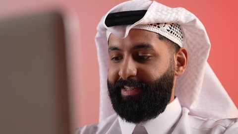 دمج الأعمال مع التقنية والتكنولوجيا الحديثة والمتطورة، شاب سعودي يقوم بإنجاز العمل عبر اللابتوب بإيماءات السعادة، بورتريه مقرب لرجل عربي خليجي قطري يرتدي الثوب التقليدي والغترة يقوم باستخدام الحاسوب المحمول، خلفية ملونة