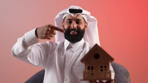 امتلاك سكن حديث والاستقرار العائلي، شاب قطري ينظر إلى الكاميرا بايماءات السعادة بشراء عقار جديد, بورتريه لرجل عربي خليجي سعودي يرتدي الثوب التقليدي والشماغ يجلس على كرسي مريح يحمل بيده مجسم بيت جديد ويشير إليه بإصبع السبابة، مفهوم الاسكان والتمليك، خلفية ملونة
