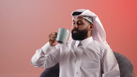 محاولة الحصول على القليل من الراحة، الاسترخاء وتناول مشروبات دافئة، شاب سعودي يقوم بالاستمتاع بكوب من القهوة الساخنة، بورتريه لرجل عربي خليجي قطري يرتدي الثوب التقليدي والغترة يجلس على كرسي ويمسك بيده فنجان من مشروبه المفضل، خلفية ملونة