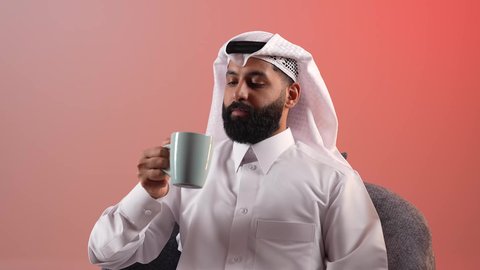 الاسترخاء وتناول مشروبات دافئة، شاب سعودي يقوم بالاستمتاع بكوب من القهوة الساخنة، بورتريه لرجل عربي خليجي قطري يرتدي الثوب التقليدي والغترة يجلس على كرسي ويمسك بيده فنجان من مشروبه المفضل، محاولة الحصول على القليل من الراحة، خلفية ملونة