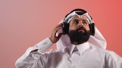 الاستماع إلى الموسيقا والبودكاست، شاب سعودي يقوم باستخدام تقنية الهيدفون، بورتريه لرجل عربي خليجي قطري يرتدي الثوب التقليدي والغترة يجلس على كرسي مريح ويضع سماعة الأذنين، دمج التقنيات والأجهزة الحديثة والتكنولوجيا بالحياة اليومية، خلفية ملونة