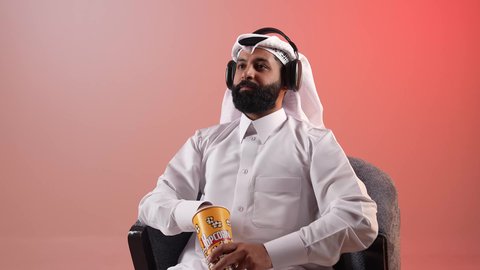 Having a nice and quiet time, listening to music and podcasts, a Saudi young man using headphones and eating popcorn, a portrait of a Qatari Gulf Arab man wearing a traditional thobe and ghutra sitting on a comfortable chair with headphones on, a colorful background