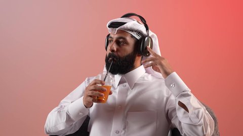 دمج التقنيات والأجهزة الحديثة والتكنولوجيا بالحياة اليومية، الاستماع إلى الموسيقا والبودكاست، شاب سعودي يقوم باستخدام تقنية الهيدفون وتناول عصير بارد، بورتريه لرجل عربي خليجي قطري يرتدي الثوب التقليدي والغترة يجلس على كرسي مريح ويضع سماعة الأذنين، خلفية ملونة