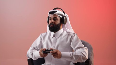 قضاء أوقات ممتعة باستخدام التقنيات الحديثة، شاب سعودي يرتدي سماعات الرأس، التفاعل والاندماج مع ألعاب الفيديو الترفيهية، بورتريه لرجل عربي خليجي قطري مبتسم يرتدي الثوب والغترة يجلس على كرسي مريح يمسك عصا التحكم بتعابير الاندماج والسرور، خلفية ملونة