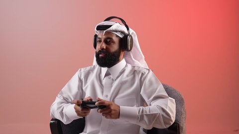قضاء أوقات ممتعة باستخدام التقنيات الحديثة، شاب سعودي يرتدي سماعات الرأس، التفاعل والاندماج مع ألعاب الفيديو الترفيهية، بورتريه لرجل عربي خليجي قطري مبتسم يرتدي الثوب والغترة يجلس على كرسي مريح يمسك عصا التحكم بتعابير الاندماج والسرور، خلفية ملونة