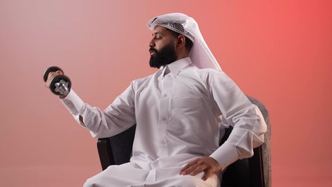 بناء الكتلة العضلية للجسم، اتباع أسلوب حياة صحي، شاب سعودي يقوم بالتدرب على رفع الأثقال والقيام بالتمارين الرياضية، بورتريه لرجل عربي خليجي قطري يرتدي الثوب التقليدي والغترة يجلس على كرسي مريح ويقوم برفع الدامبلز، خلفية ملونة