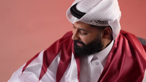 إحياء ذكرى تأسيس الدولة، الاحتفال باليوم الوطني القطري 18 ديسمبر، بورتريه مقرب لرجل عربي خليجي قطري يجلس على كرسي مريح يضع علم قطرعلى كتفيه بايماءات الفخروالاعتزاز، رموز وثقافات وطنية, حب الوطن والانتماء إليه، خلفية ملونة