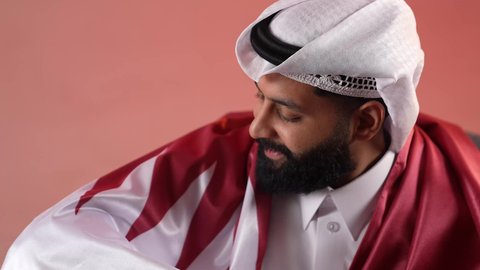 Commemorating the founding of the state, celebrating Qatar National Day, December 18, a close-up portrait of a Qatari Gulf Arab man sitting on a comfortable chair with the Qatari flag on his shoulders, gestures of pride and honor, national symbols and cultures, love of the homeland and belonging to it, colored background