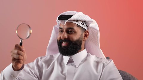تكبير وتوضيح الرؤية، شاب سعودي يقوم بعمليات البحث والتدقيق، مضاعفة الأحجام ومحاولة إيجاد الأشياء الصغيرة، بورتريه مقرب لرجل عربي خليجي قطري يرتدي الثوب والغترة يجلس على كرسي يحاول اكتشاف شيء ما باستخدام العدسة المكبرة، إيماءات الاندماج والسرور، خلفية ملونة