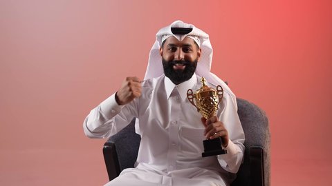 ايماءات النجاح وتحقيق الانجازات، شاب سعودي يقوم بتشجيع الفرق والمنتخبات الرياضية, بورتريه لرجل عربي خليجي قطري مبتسم يرتدي الثوب التقليدي والغترة يجلس على كرسي يحمل بيده كأس البطولة ويرفع قبضة يده بتعابير الفوز, خلفية ملونة