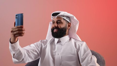 Preserving happy moments and souvenir photos, modern and advanced technology, a bearded Saudi young man using a mobile phone, a portrait of a Qatari Gulf Arab man sitting on a comfortable chair taking a selfie with his mobile phone, gesturing with pleasure and happiness, a colorful background