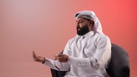 تحديد شيء معين، شاب سعودي يجلس على كرسي مريح ينظر إلى الكاميرا بإيماءات السرور والسعادة، بورتريه لرجل عربي خليجي قطري مبتسم يرتدي الثوب التقليدي والغترة يفتح كفه للأعلى ويشير إلى عنصر ما, حمل عنصر هولوجرامي، خلفيه ملونة
