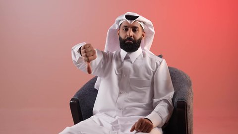 Feeling of dissatisfaction and rejection, negative evaluation, portrait of a Qatari Gulf Arab man wearing a traditional thobe and ghutra looking at the camera, a Saudi young man sitting on a comfortable chair giving a thumbs down gesture of annoyance, anxiety and failure in something, colorful background