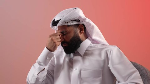 Headache and chronic sinusitis, a young Saudi man sitting on a comfortable chair feeling the need for regular checkups and follow-ups, illness, cold and flu, portrait of a Qatari Gulf Arab man wearing traditional thobe and ghutra putting his hand on his nose with an expression of pain, colored background