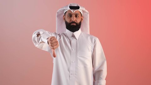 الرفض والتقييم السلبي، بورتريه لرجل عربي خليجي قطري يرتدي الثوب التقليدي والغترة ينظر الى الكاميرا, شاب سعودي يقوم بالإشارة بإصبع الابهام للأسفل بإيماءات الإنزعاج, القلق والفشل في أمر ما, الشعور بعدم القبول والرضا, خلفية ملونة