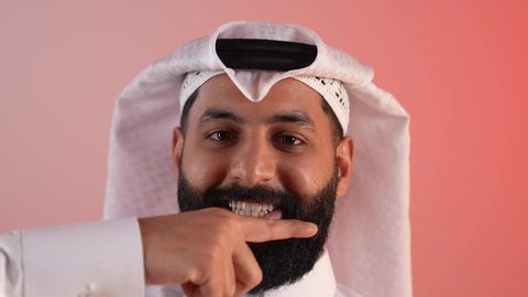 النظر إلى الكاميرا بتعابير السرور، تحديد موقع شيء معين، شاب سعودي يقوم بالاشارة إلى شخص ما، بورتريه مقرب لرجل عربي خليجي قطري مبتسم يرتدي الثوب التقليدي والغتره يشير باصبع السبابة إلى الجانب، مفهوم الأناقة والاهتمام بالمظهر الخارجي، خلفيه ملونة