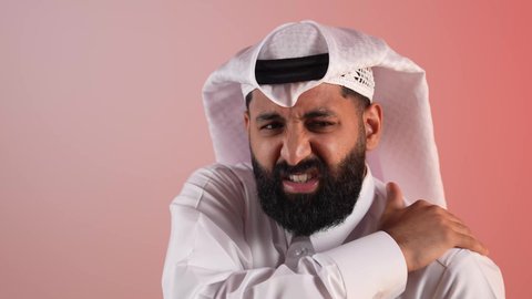 التهاب المفاصل والعضلات، آلام العظام والعمود الفقري، الحاجة للذهاب إلى الطبيب والمعاينة، شاب سعودي يشعر بضرورة الاسترخاء وأخذ قسط من الراحة، بورتريه مقرب لرجل عربي خليجي قطري يرتدي الثوب والغتره يضع يده على كتفه بإيماءات التألم، خلفية ملونة