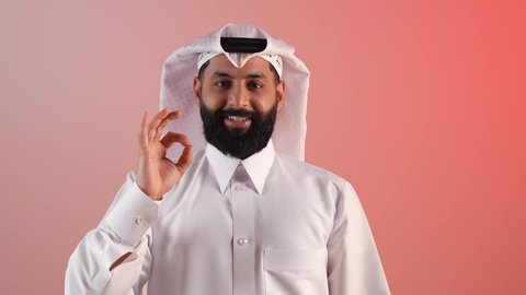 رفع اليد بعلامات الجودة والقبول، التعبير عن الإعجاب والتقدير، الامتياز والعلامة الكاملة، الشعور بالرضا والاستحسان، بورتريه مقرب لرجل عربي خليجي قطري مبتسم يرتدي الثوب التقليدي والغتره، شاب سعودي ينظر إلى الكاميرا بإيماءات السعادة والسرور، خلفية ملونة