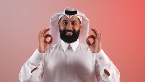 رفع اليد بعلامات الجودة والقبول، التعبير عن الإعجاب والتقدير، الامتياز والعلامة الكاملة، الشعور بالرضا والاستحسان، بورتريه مقرب لرجل عربي خليجي قطري مبتسم يرتدي الثوب التقليدي والغتره، شاب سعودي ينظر إلى الكاميرا بإيماءات السعادة والسرور، خلفية ملونة