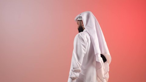 الاهتمام بالمظهر الخارجي، مفهوم الأناقة والوسامة، بورتريه من الخلف لرجل عربي خليجي سعودي يرتدي الثوب التقليدي والغترة، شاب قطري ملتحي يقوم باللتفات والنظر إلى الكاميرا بإيماءات الثقة بالنفس، خلفية ملونة