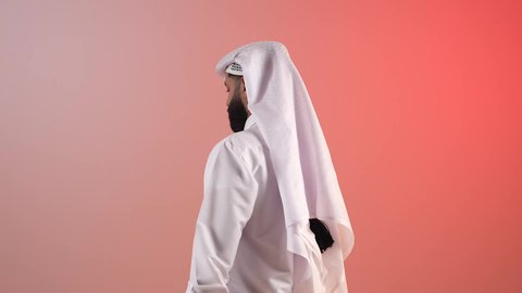 Concern for outward appearance, concept of elegance and handsomeness, portrait from the back of a Saudi Arabian Gulf man wearing traditional thobe and ghutra, a bearded Qatari young man turning and looking at the camera with gestures of self-confidence, colorful background