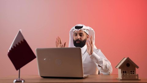 Holding online meetings via video calls, using a laptop at work, answering and responding to questions, inquiries and calls, a portrait of a Qatari Gulf Arab man wearing a traditional thobe and ghutra sitting behind a desk in front of him with a laptop, the Qatari flag and a house model, a colorful background