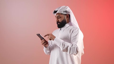 التقنية الحديثة والمتطورة، التواصل مع الأهل والأصدقاء، شاب سعودي ملتحي يقوم باستخدام الهاتف المحمول، بورتريه لرجل عربي خليجي قطري يقوم بتصفح مواقع التواصل الاجتماعي عبر الجوال بإيماءات السرور والسعادة، خلفية ملونة