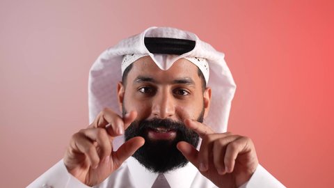 مفهوم الوسامة والرجولة والقوة، الاهتمام بالأناقة والمظهر الخارجي، بورتريه مقرب لرجل عربي خليجي قطري  يرتدي الثوب التقليدي والغترة يقوم بتعديل شاربه، شاب سعودي ينظر إلى الكاميرا بإيماءات السرور والثقة في النفس، خلفية ملونة