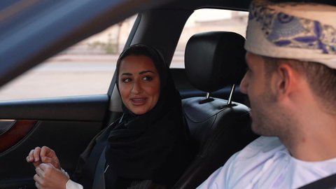 Family group trip in the car, strolling in the streets of the Sultanate of Oman with the family, chatting and conversations on the road, an Arab Gulf Omani man wearing a dishdasha and turban sitting with his wife inside the car, gestures of happiness and pleasure, spending a good time with the family, wearing a seat belt