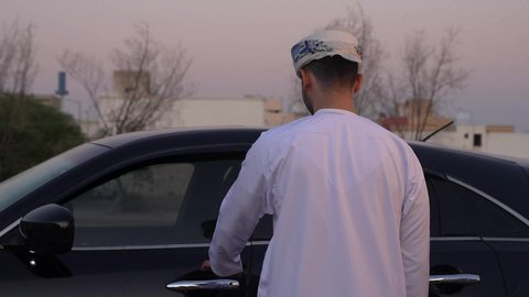 تعابير السعادة والسرور, الاستعداد لركوب السيارة، مفهوم القيادة الآمنة, رجل عربي خليجي عماني يرتدي الدشداشة و العمامة يفتح باب السيارة الامامي، خدمة النقل و التوصيل في سلطنة عمان