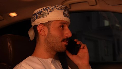 اجراء مكالمة هاتفية عبر الهاتف المحمول، استخدام جهاز تقني حديث ومتطور، خدمة النقل و التوصيل في سلطنة عمان، الالتزام بوضع حزام الأمان, مفهوم القيادة الآمنة, سائق عربي خليجي عماني يرتدي الدشداشة و العمامة  يقود السيارة بتعابير السعادة والسرور