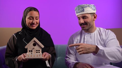الاشارة الى المنزل، الاستقرار العائلي وامتلاك سكن حديث، مفهوم الاسكان وتمليك، رجل عربي خليجي عماني يرتدي الدشداشة والعمامة يشير باصبع السبابة الى الجانب، إمرأة تمسك مجسم منزل صغير، تعابير السرور والسعادة بشراء عقار جديد, خلفية بنفسجية