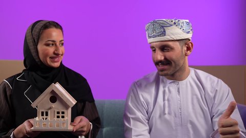 ايماءات الاعجاب والقبول، الاستقرار العائلي وامتلاك سكن حديث، مفهوم الاسكان وتمليك، رجل عربي خليجي عماني يرتدي الدشداشة والعمامة يرفع اصبع الابهام لاعلى، إمرأة تمسك مجسم منزل صغير، تعابير السرور والسعادة بشراء عقار جديد, خلفية بنفسجية