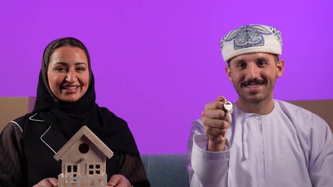الاستقرار العائلي وامتلاك سكن حديث، صناديق وحاويات كرتونية, مفهوم الاسكان وتمليك، رجل عربي خليجي عماني يرتدي الدشداشة والعمامة يحمل بيده مفتاح بيت جديد يقف جانب زوجته، إمرأة تمسك مجسم منزل صغير، ايماءات السرور والسعادة بشراء عقار جديد, خلفية بنفسجية