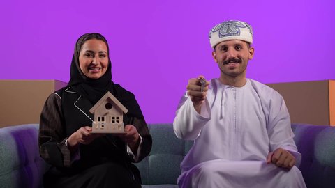 الاستقرار العائلي وامتلاك سكن حديث، صناديق وحاويات كرتونية, مفهوم الاسكان وتمليك، رجل عربي خليجي عماني يرتدي الدشداشة والعمامة يحمل بيده مفتاح بيت جديد يقف جانب زوجته، إمرأة تمسك مجسم منزل صغير، ايماءات السرور والسعادة بشراء عقار جديد, خلفية بنفسجية