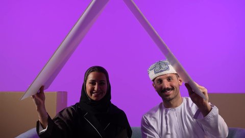 Looking at the camera, gestures of happiness for buying a new property, owning a modern home and family stability, cardboard boxes and containers, the concept of housing and ownership, an Arab Gulf Omani man wearing a dishdasha and turban holding a model of the roof of the house sitting next to his wife, purple background