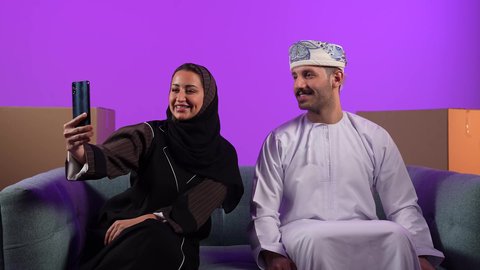 الانتقال والاسكان وتمليك، صناديق وحاويات كرتونية, إمرأة عربية خليجية عمانية ترتدي الحجاب والعباءة تقوم بالتقاط سيلفي باستخدام الهاتف المحمول مع زوجها، ايماءات السعادة بشراء عقار جديد, امتلاك سكن حديث والاستقرار العائلي، خلفية بنفسجية