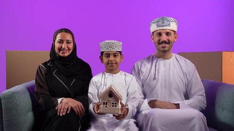 امتلاك سكن حديث والاستقرار العائلي، صناديق وحاويات كرتونية, مفهوم الاسكان وتمليك، ولد عربي خليجي عماني يرتدي الدشداشة والكمة يحمل بيده مجسم بيت صغير يجلس امام عائلته، ايماءات السرور والسعادة بشراء عقار جديد, خلفية بنفسجية