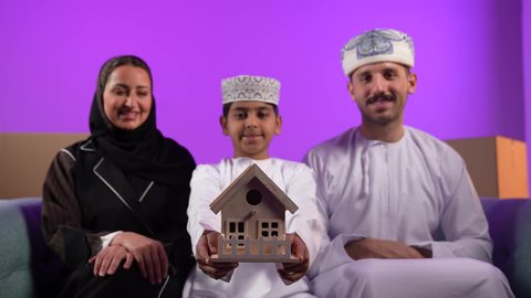 امتلاك سكن حديث والاستقرار العائلي، صناديق وحاويات كرتونية, مفهوم الاسكان وتمليك، ولد عربي خليجي عماني يرتدي الدشداشة والكمة يحمل بيده مجسم بيت صغير يجلس امام عائلته، ايماءات السرور والسعادة بشراء عقار جديد, خلفية بنفسجية