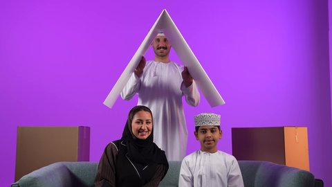 Cardboard boxes and containers, housing and ownership concept, an Omani Gulf Arab man wearing a dishdasha and turban holding a model of the house roof in front of his family, an Omani wife sitting next to her son looking at the camera with gestures expressing happiness at buying a new property, owning a modern home and family stability, purple background