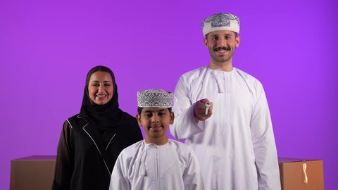 الاستقرار العائلي وامتلاك سكن حديث، صناديق وحاويات كرتونية, مفهوم الاسكان وتمليك، رجل عربي خليجي عماني يرتدي الدشداشة والعمامة يحمل بيده مفتاح بيت جديد يقف امام عائلته، ايماءات السرور والسعادة بشراء عقار جديد, خلفية بنفسجية