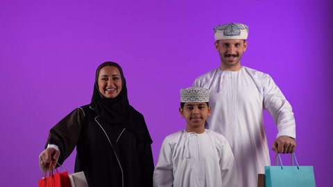 متعة التسوق مع الأبناء والعائلة في المولات، عائلة عربية خليجية عمانية تحمل بيدها اكياس تسوق ملونة، شراء واقتناء المستلزمات والهدايا من المولات، حسومات وعروض الجمعة البيضاء في سلطنة عمان، خلفية بنفسجية