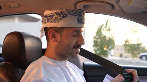 خدمة النقل و التوصيل في سلطنة عمان، الالتزام بوضع حزام الأمان, مفهوم القيادة الآمنة, سائق عربي خليجي عماني يرتدي الدشداشة و العمامة يمسك المقود بيده و يقود السيارة بتعابير السعادة والسرور