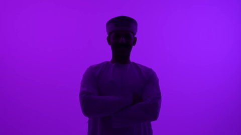 مفهوم الرجولة والوسامة، الوقوف بأيدي متقاطعة بإيماءات الثقة بالنفس، تصوير ظلي مقرب لشاب عربي خليجي عماني يرتدي الدشداشه والعمامة ينظر إلى الكاميرا، الاهتمام بالمظهر الخارجي والأناقة، خلفية بنفسجية، سالوييت