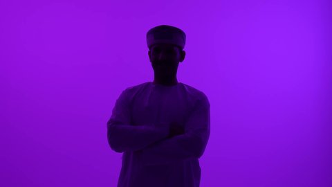 الوقوف بأيدي متقاطعة بإيماءات الثقة بالنفس، مفهوم الرجولة والوسامة،  تصوير ظلي مقرب لشاب عربي خليجي عماني يرتدي الدشداشه والعمامة ينظر إلى الكاميرا، الاهتمام بالمظهر الخارجي والأناقة، خلفية بنفسجية، سالوييت