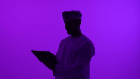 ادارة العمل عن بعد، استخدام جهاز تقني حديث ومتطور, تصوير ظلي مقرب لرجل عربي خليجي عماني يرتدي الدشداشة والعمامة يعمل على جهاز التابلت, إنجاز المهام المطلوبة عبر الجهاز اللوحي، خلفية بنفسجية، سالوييت