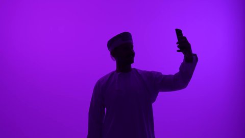 الدردشة مع الأهل والأصدقاء عبر الانترنت، استخدام الأجهزة الحديثة والمتطورة، إجراء مكالمة فيديو عبر وسائل التواصل الاجتماعي، تصوير ظلي مقرب لشاب عربي خليجي عماني يرتدي الدشداشة والعمامة يقوم بالتقاط صورة سيلفي، خلفيه بنفسجية، سالوييت