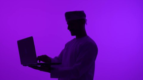 استخدام جهاز تكنولوجي متطور، عرض تطبيقات وخدمات جهاز الحاسوب، تصوير ظلي مقرب لرجل عربي خليجي عماني يرتدي الدشداشة و العمامة يحمل بيده جهاز الحاسوب المحمول، انجاز مهام العمل عن بعد، خلفية بنفسجية، سالوييت