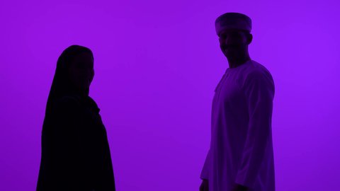 الاهتمام بالاناقة والمظهر الخارجي، الوقوف باستقامة والنظر الى الكاميرا، تصوير ظلي مقرب لرجل عربي خليجي عماني يرتدي الدشداشة والعمامة يقف بجانب زوجته، خلفية بنفسجية، سالوييت