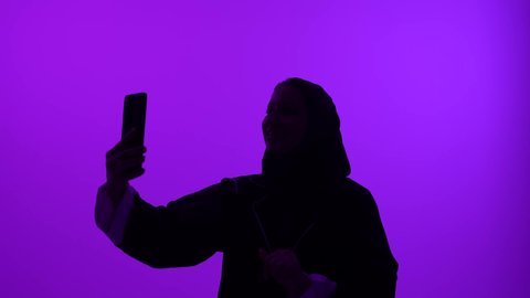 Chatting with family and friends online, Omani woman using modern and advanced devices, making a video call via social media, close-up silhouette of a young Saudi Arabian Gulf woman wearing an abaya and hijab taking a selfie, purple background, silhouette
